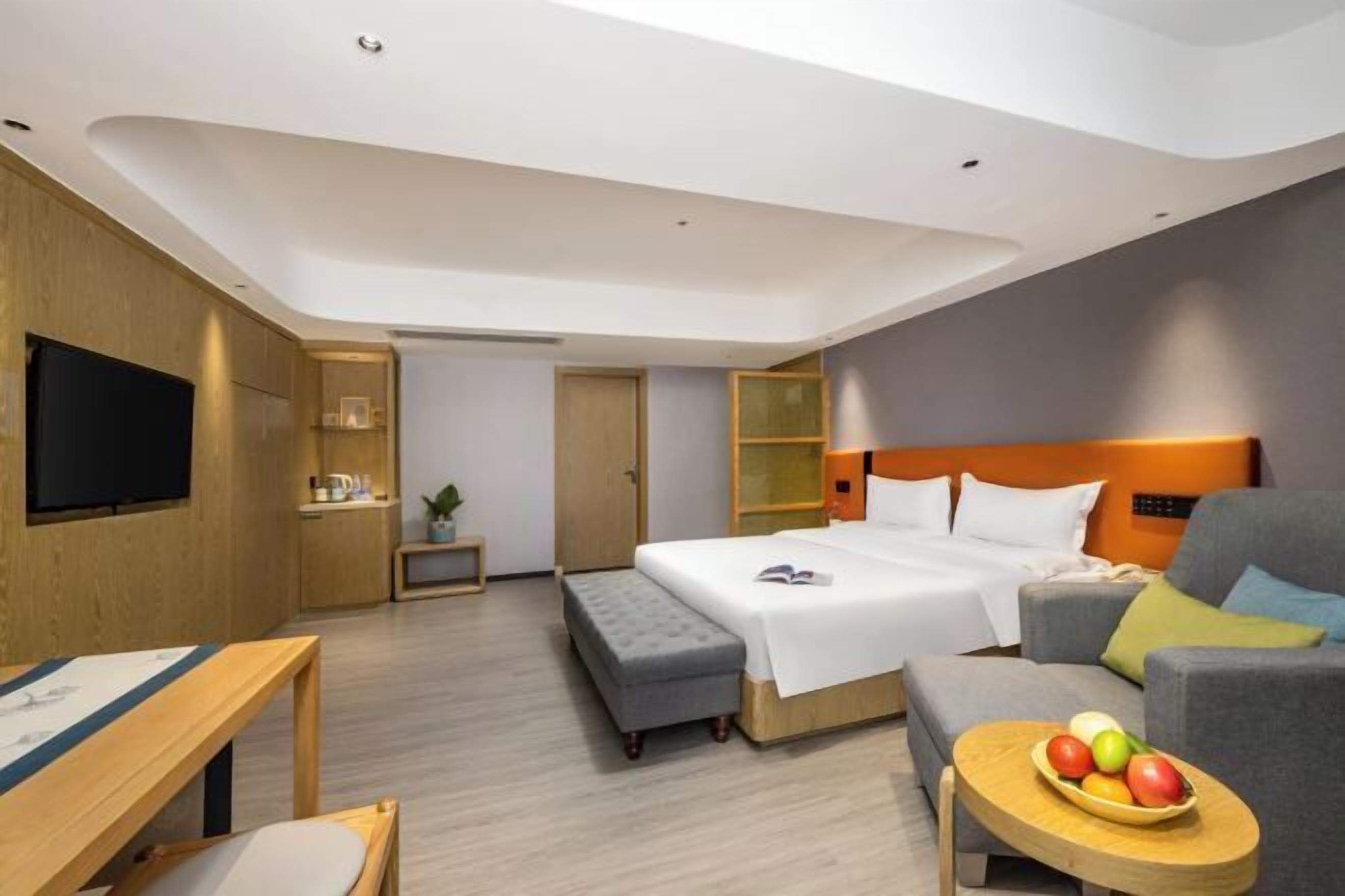 A modern hotel room with a bed, a sofa, a TV, a small table with fruit, and a bench.