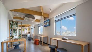 Un interior moderno con mesas y taburetes de madera, ventanas amplias con vista a la ciudad y un techo con vigas a la vista.