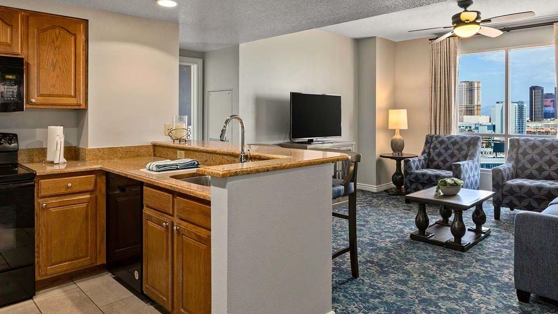 A hotel room with a kitchenette, a flat-screen TV, a seating area with a view of the city, and a ceiling fan.