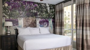 A hotel room with a large bed, a nightstand, a lamp, and a view of a building through the window.