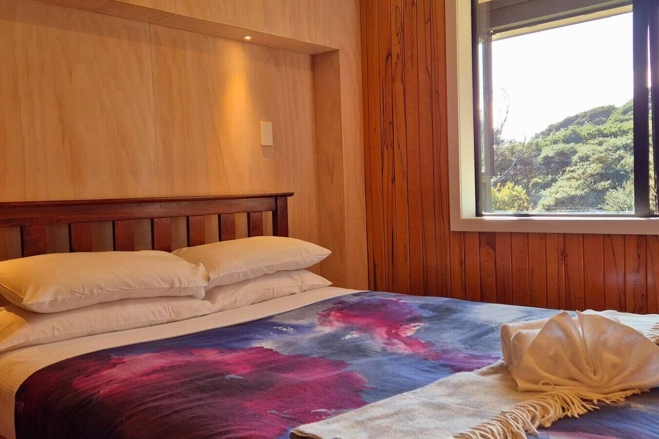 A bed with a galaxy-themed bedspread, wooden headboard, and a window with a view of greenery.