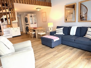A modern living room with a sofa, ottoman, dining table, and chairs. There is a kitchen area with cabinets and a stove.