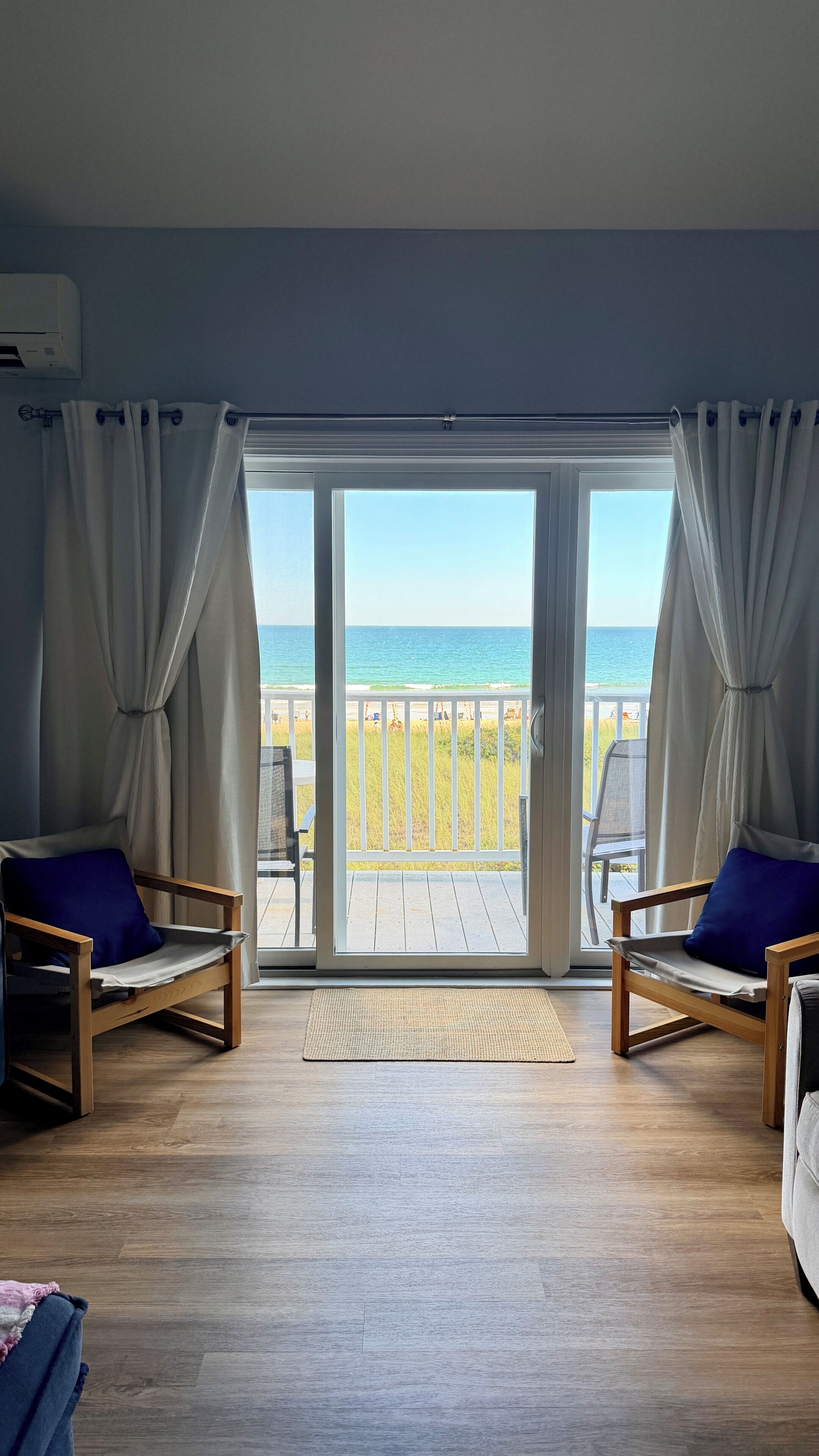 Une pièce dotée d’un plancher en bois, de deux chaises en bois avec des coussins bleus, et d’une grande porte-fenêtre coulissante qui donne sur un balcon offrant une vue sur la plage et l’océan.