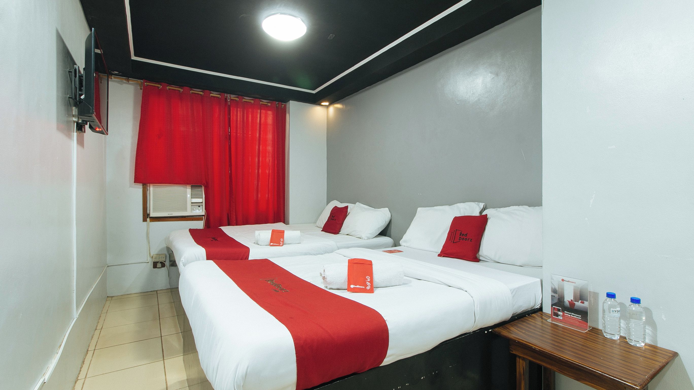Hotellihuone, jossa on kaksi sänkyä, punavalkoiset vuodevaatteet, puinen yöpöytä, jossa on vesipulloja, ja punainen verho.