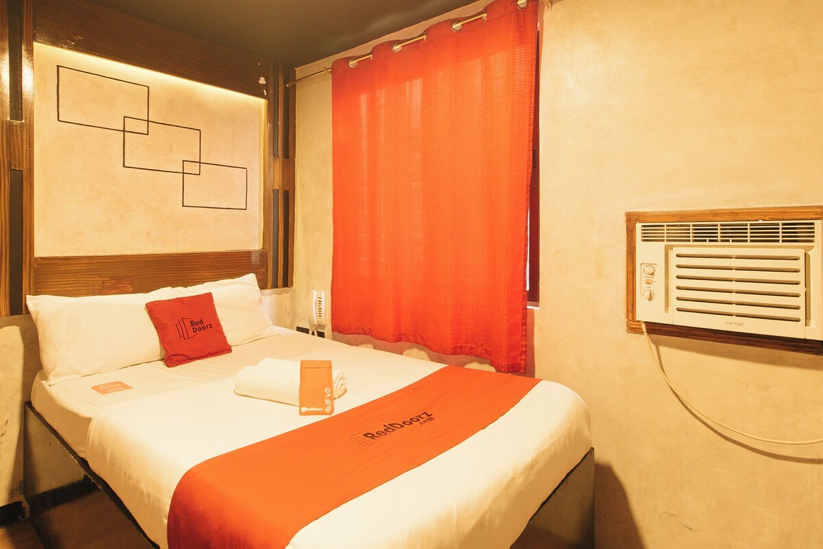 Habitación de hotel con cama, una señaleta roja en la puerta, ventana con persianas y un aire acondicionado instalado en la pared.