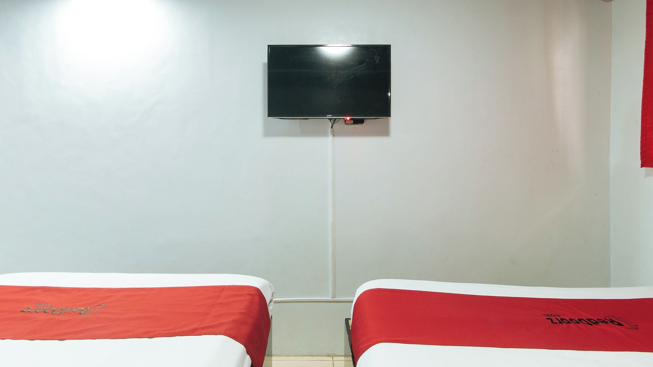 A hotel room with two beds, each with a red headboard and a white mattress cover. A flat-screen TV is mounted on the wall above the beds.