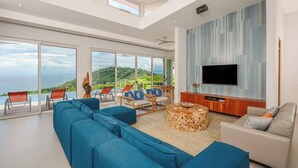 Ein modernes Wohnzimmer mit großem Fernseher, bequemen Sitzmöglichkeiten und einem schönen Meerblick.