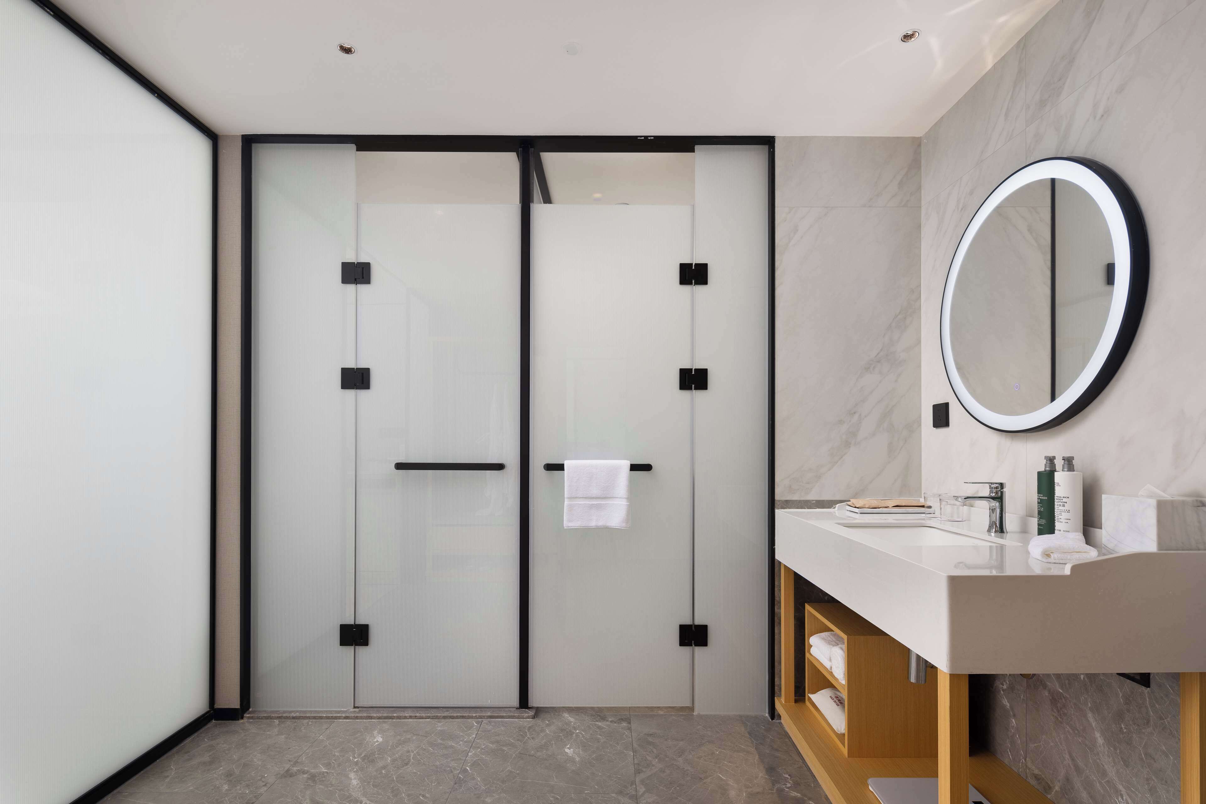 A modern bathroom with a glass door, a round mirror, and a white countertop.