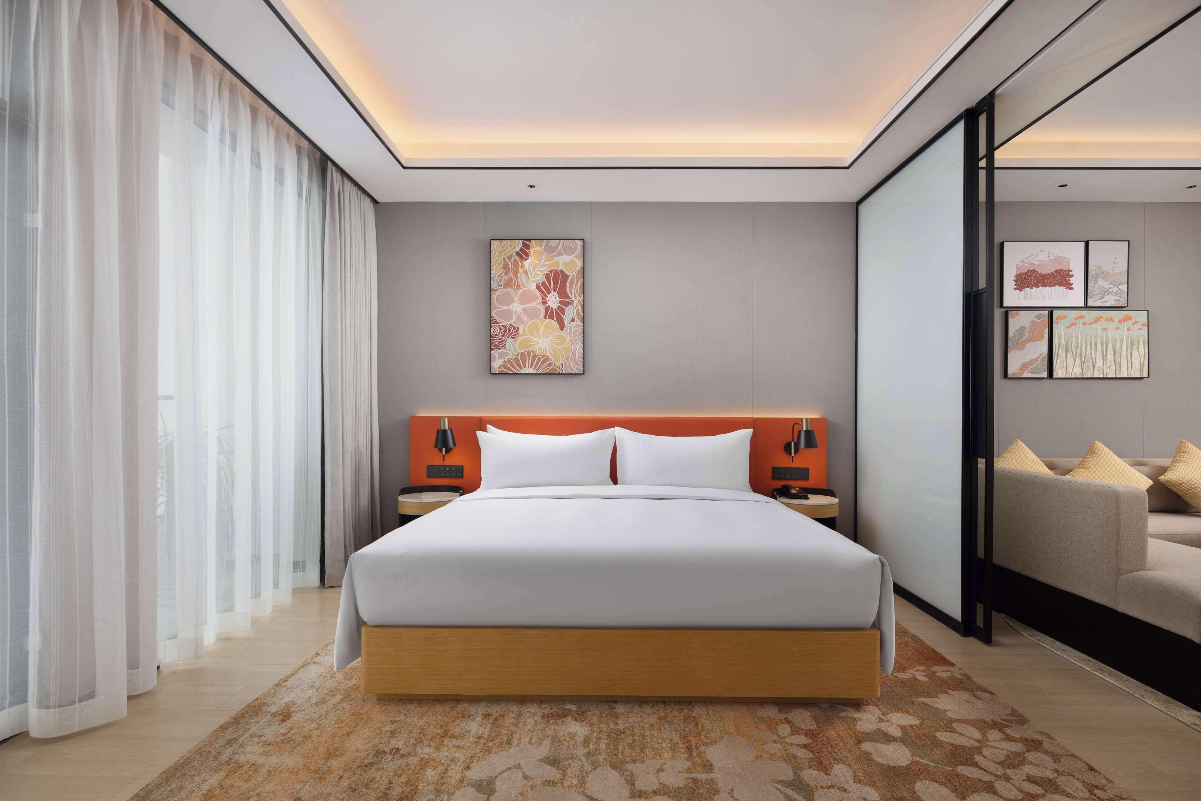 A modern hotel room with a large bed, a nightstand, a wall-mounted lamp, and a painting.