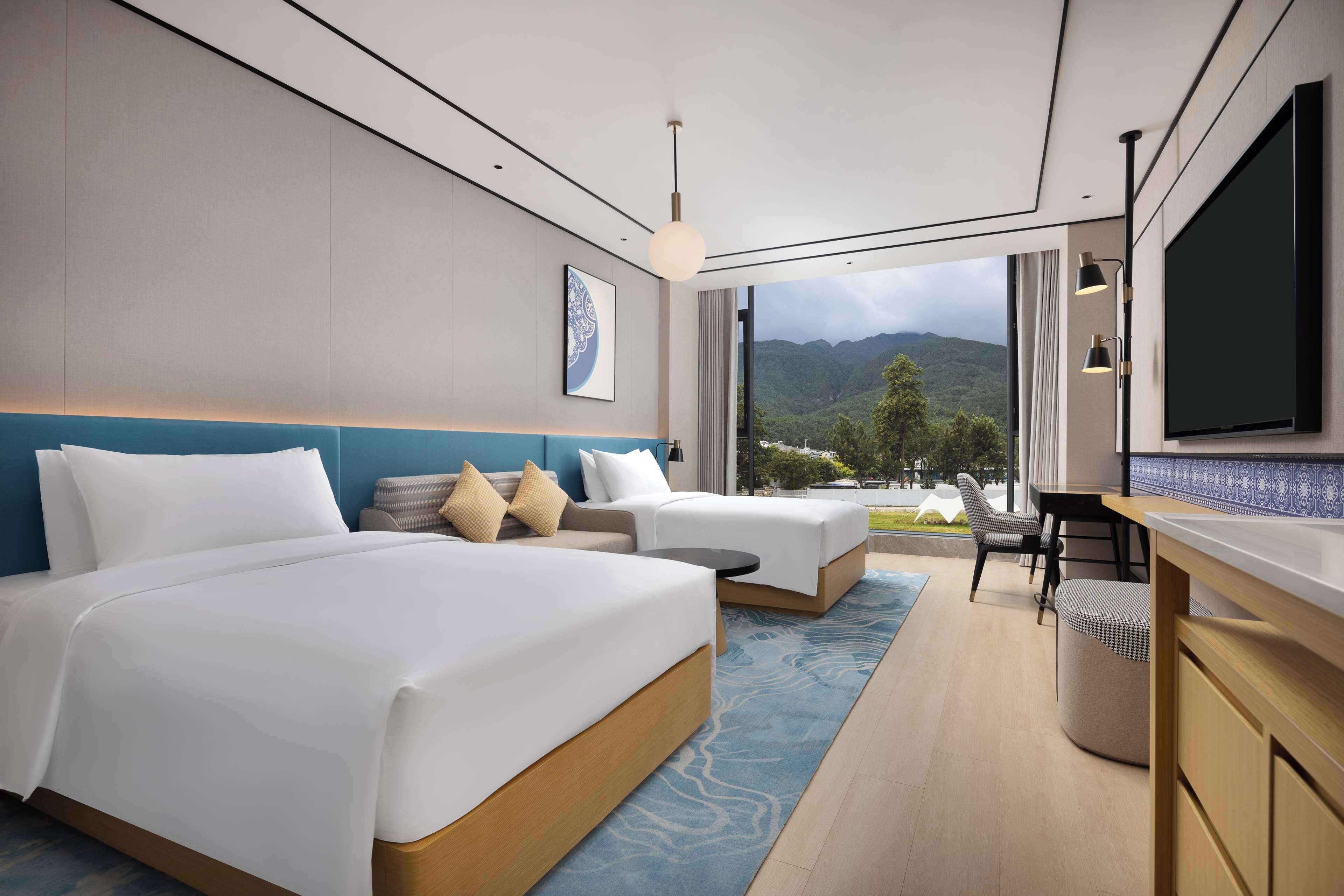 A modern hotel room with two beds, a desk, and a view of mountains.