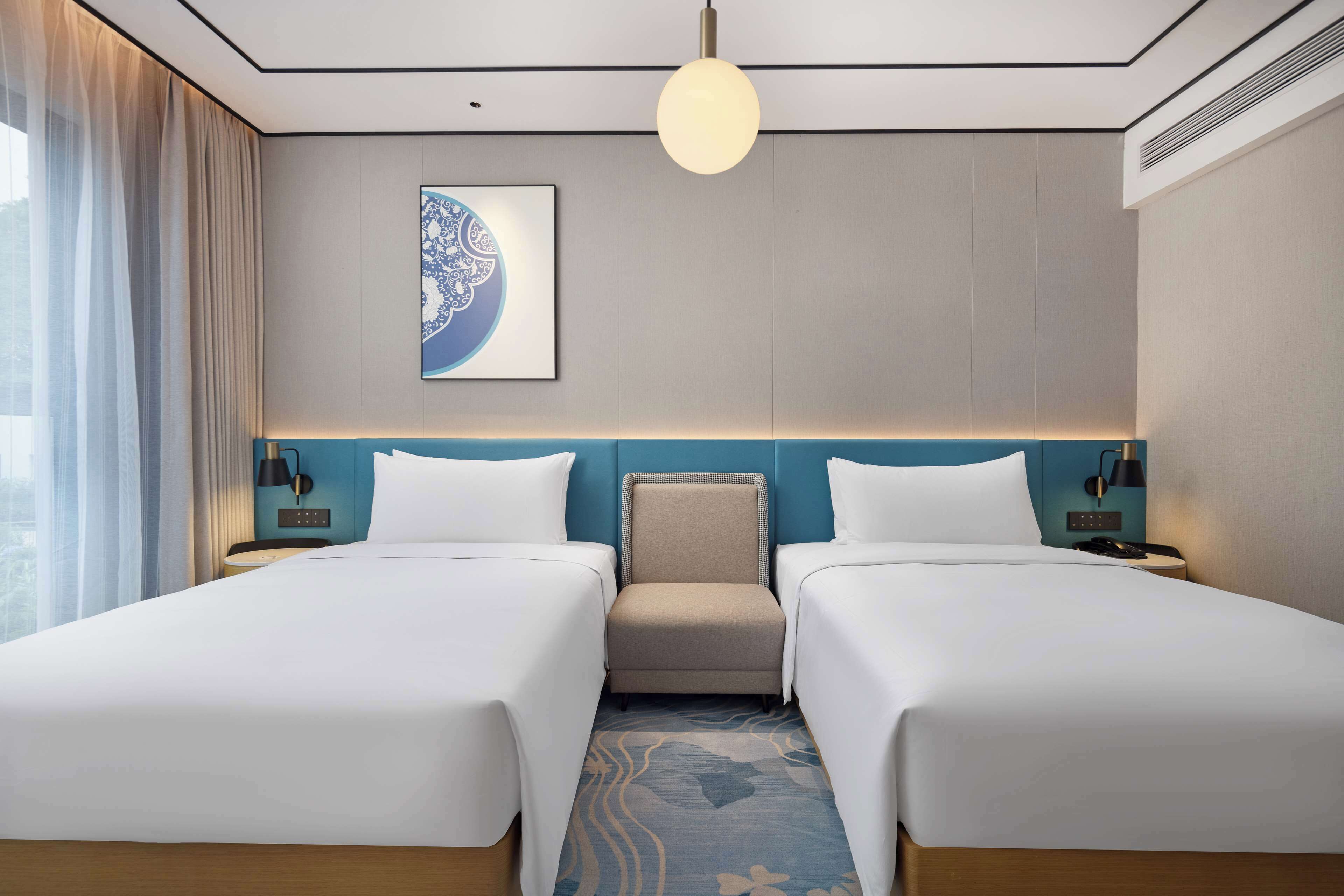 A hotel room with two beds, a headboard, a wall-mounted artwork, a ceiling light, and a patterned rug.