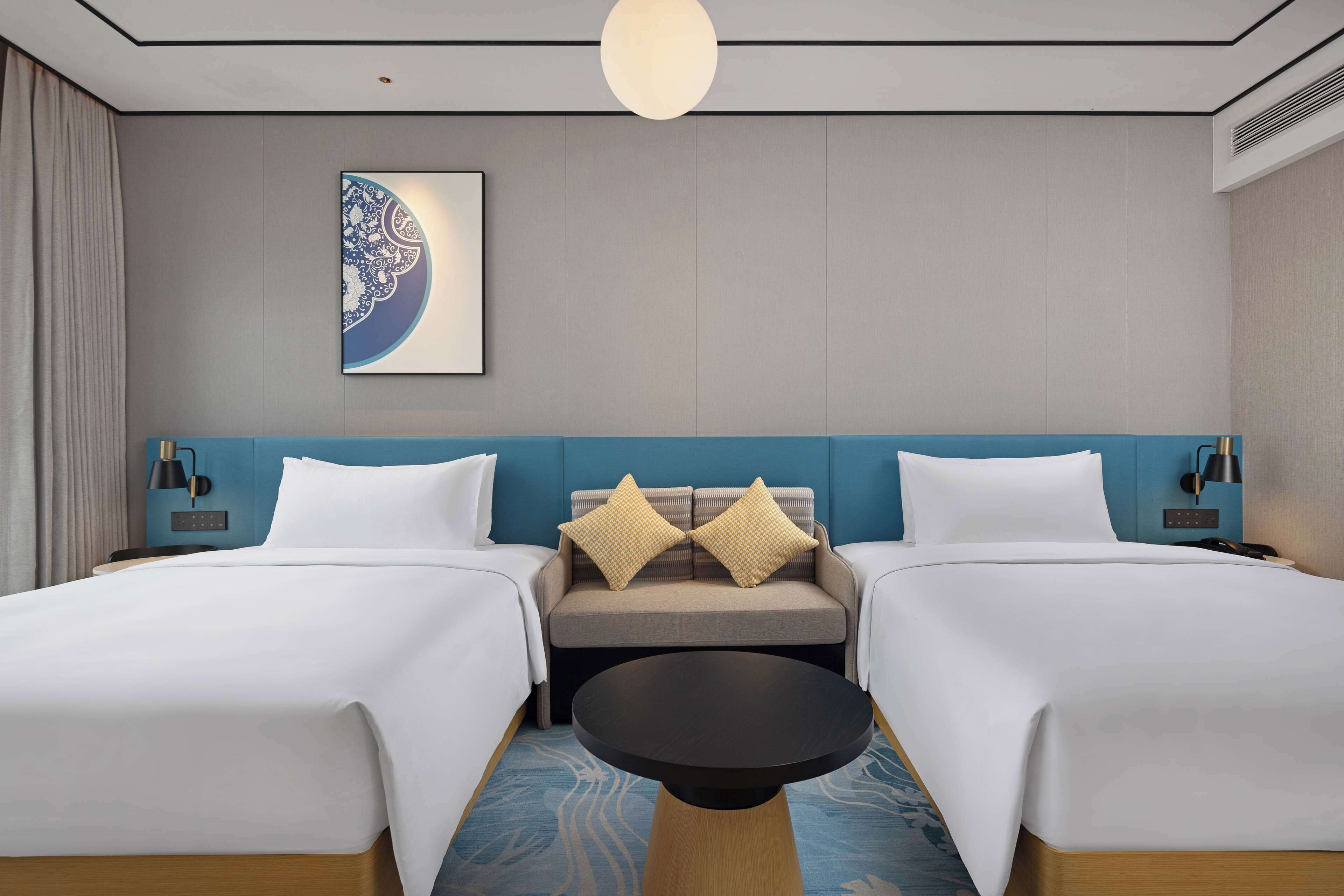 A hotel room with two beds, a sofa, a round table, and a wall-mounted artwork.