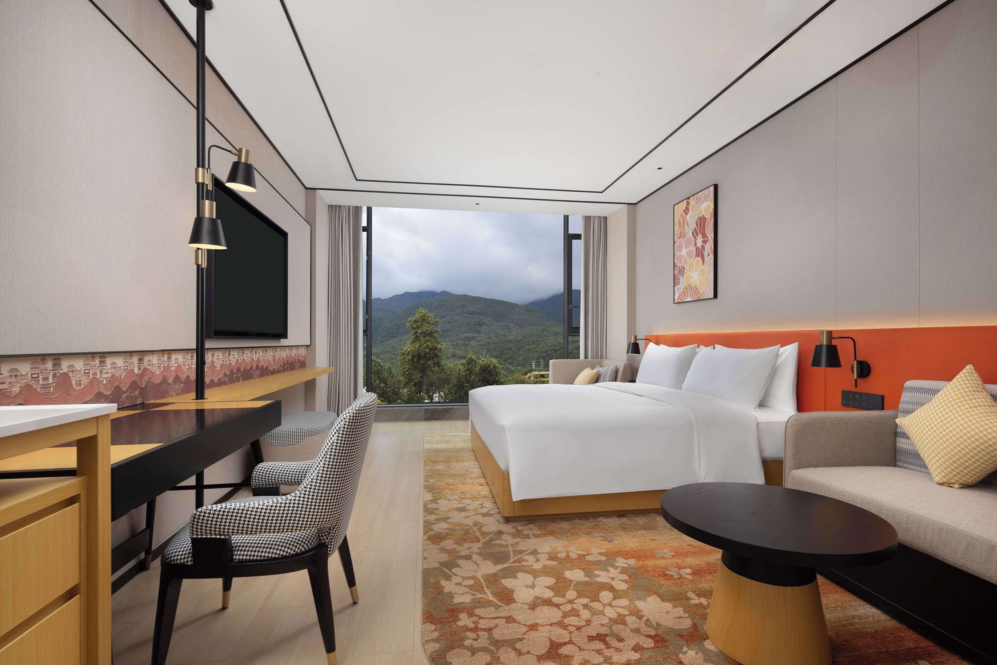 Une chambre d’hôtel moderne, dotée d’un grand lit, d’un bureau, d’une chaise, d’une petite table et offrant une vue sur les montagnes par une grande fenêtre.