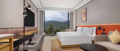 A modern hotel room with a large bed, a desk, a chair, a small table, and a view of mountains through a large window.