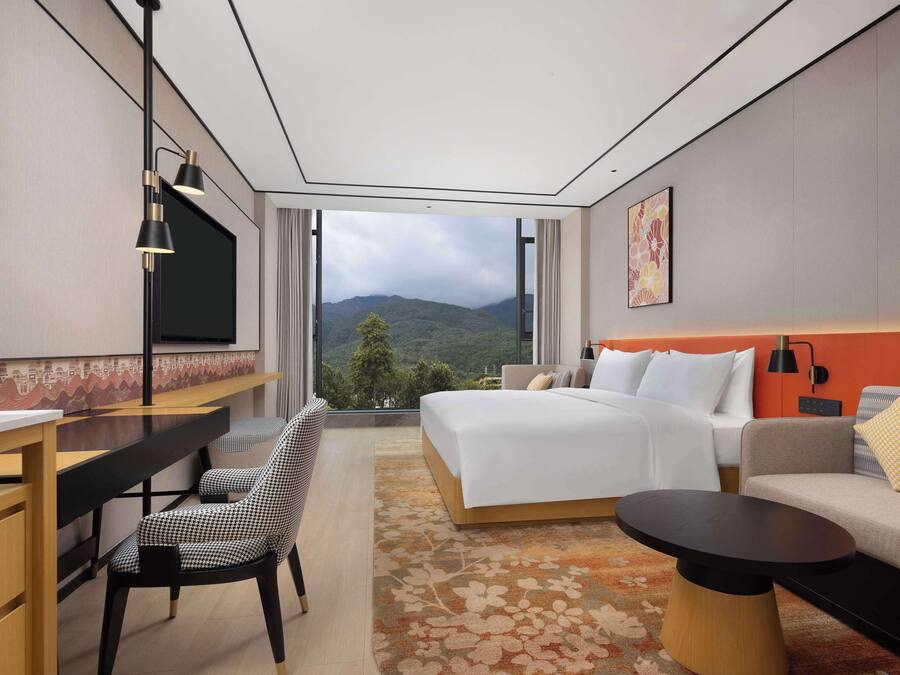 Une chambre d’hôtel moderne, dotée d’un grand lit, d’un bureau, d’une chaise, d’une petite table et offrant une vue sur les montagnes par une grande fenêtre.
