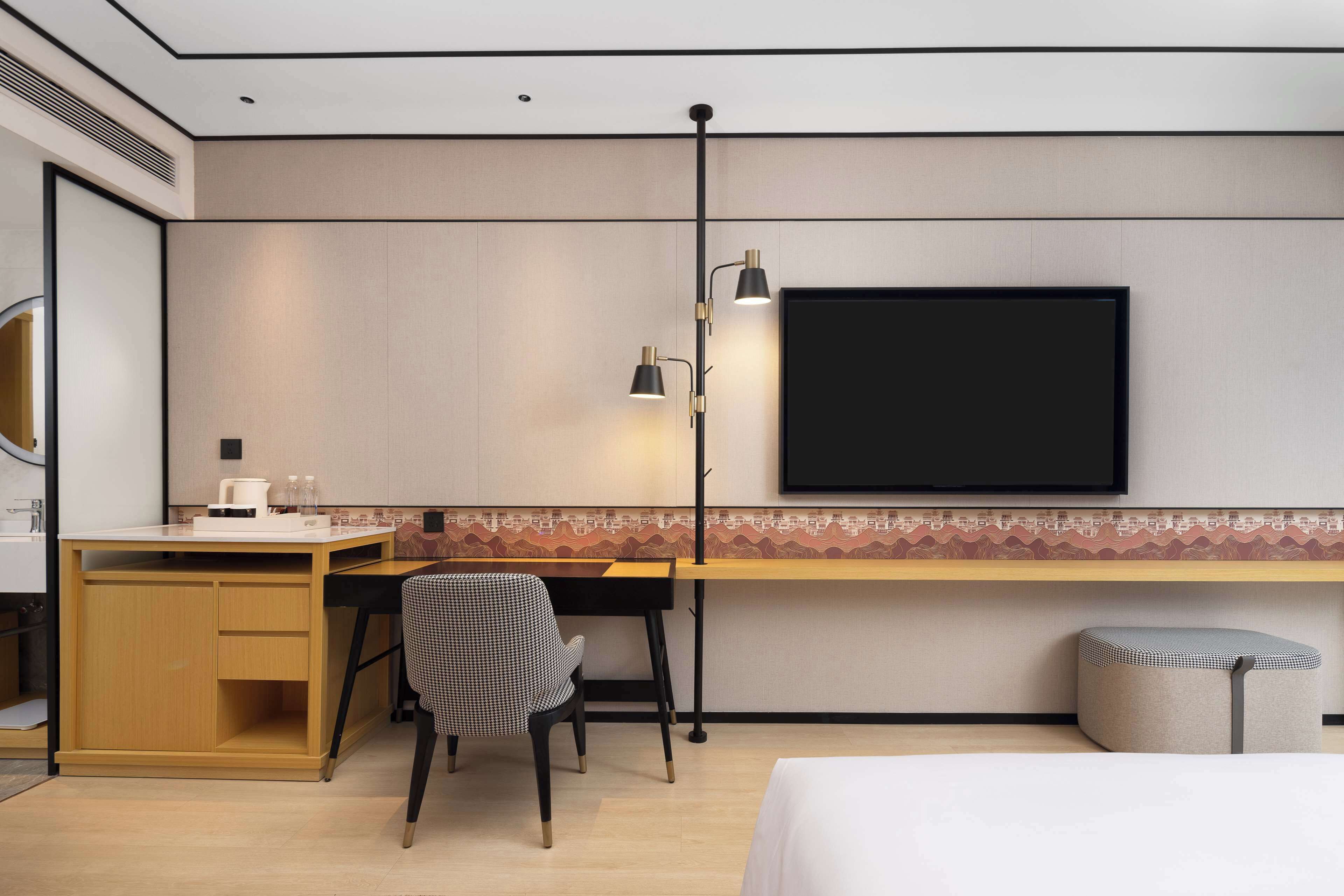 A modern hotel room with a wooden desk, a black chair, a flat-screen TV mounted on the wall, and a small ottoman.
