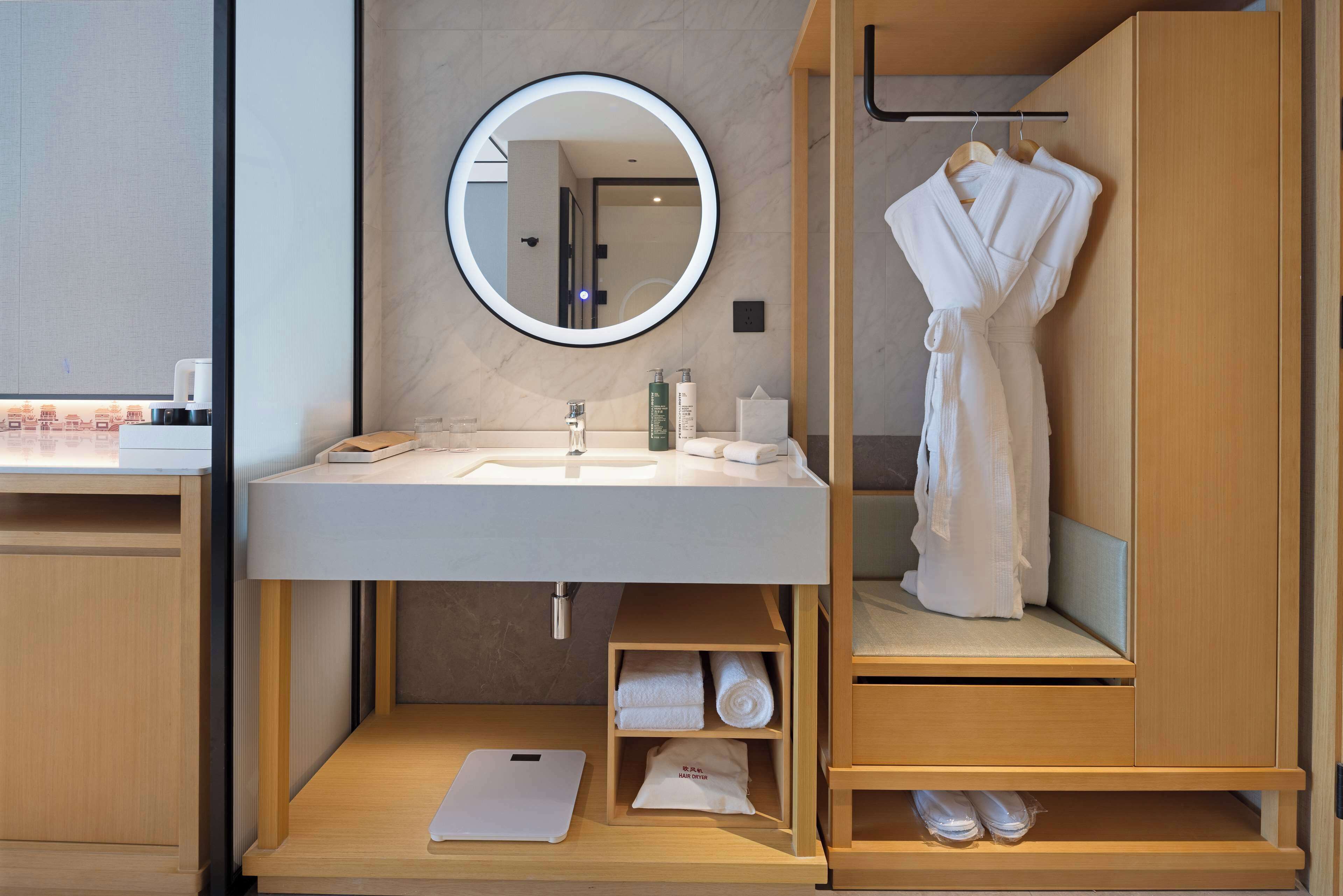 Un baño moderno con un espejo redondo, lavamanos blanco y tocador de madera. Hay toallas y una bata colgadas en la pared, y un estante con artículos de aseo.