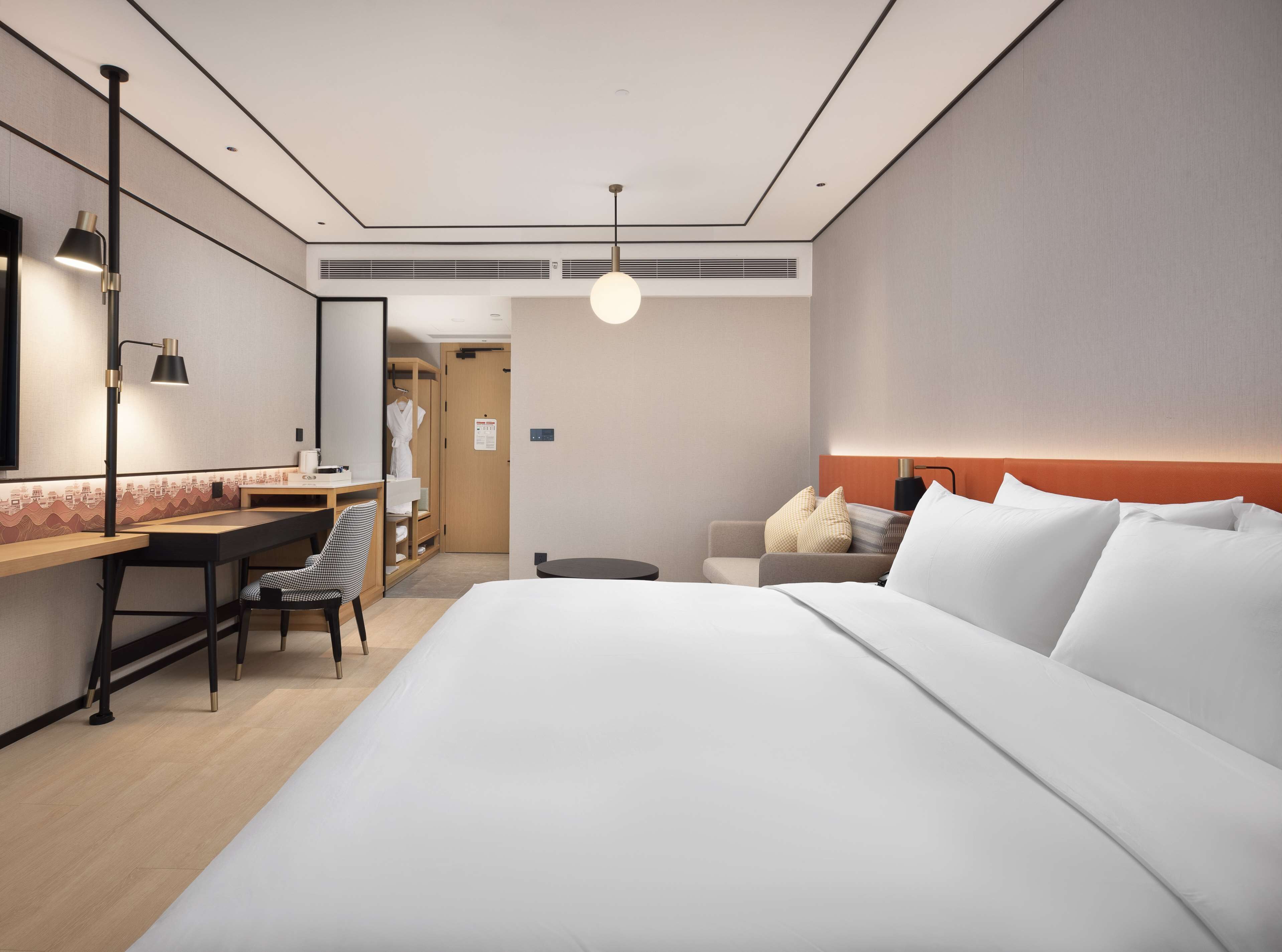 A modern hotel room with a large bed, a desk with a chair, a wardrobe, and a bedside table.
