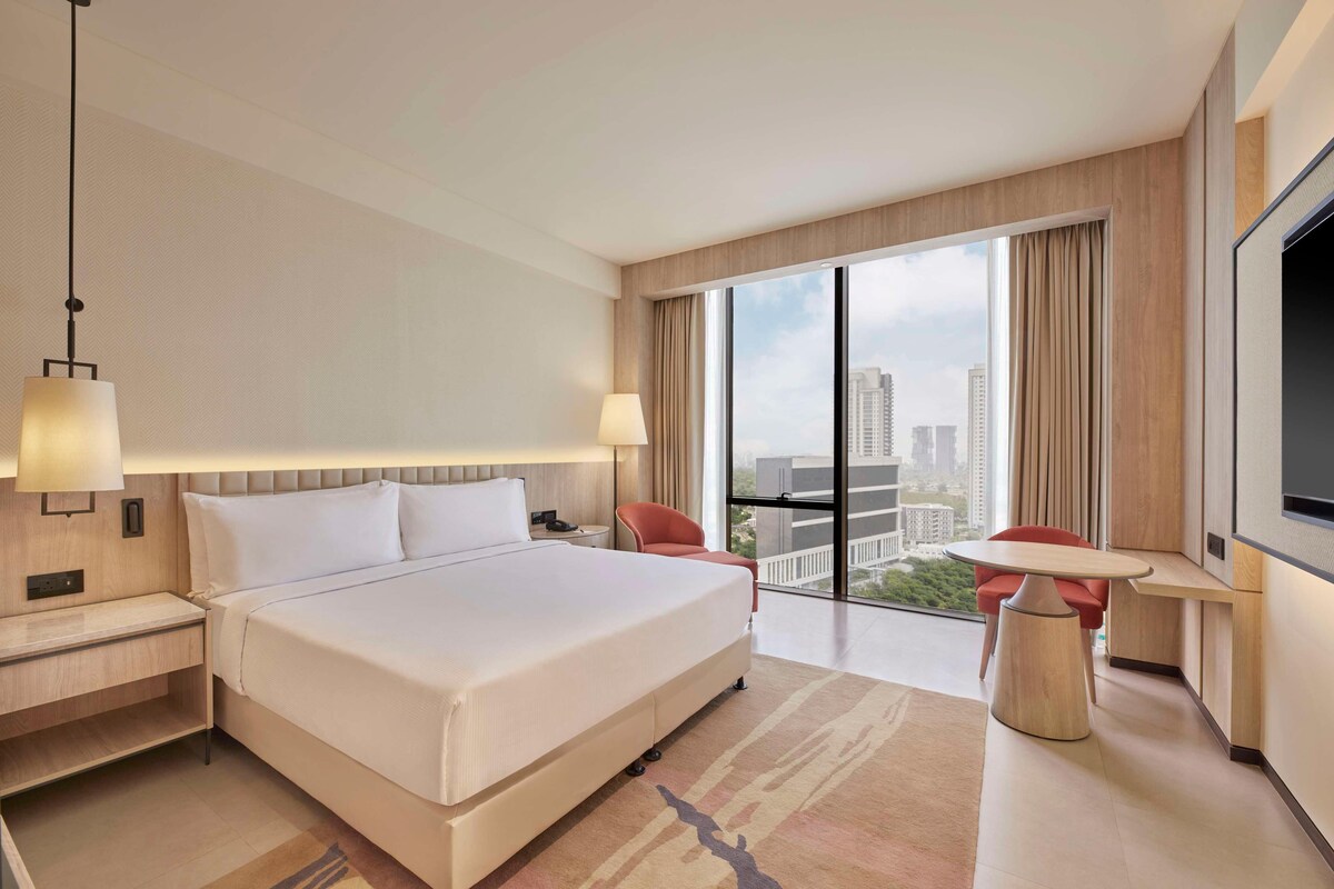 Une chambre d’hôtel dotée d’un grand lit, d’un bureau avec une chaise, d’une télévision et offrant une vue sur la ville grâce à de grandes fenêtres.