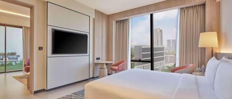 A hotel room with a large bed, a flat-screen TV mounted on the wall, a desk with a chair, and a view of the city through large windows.