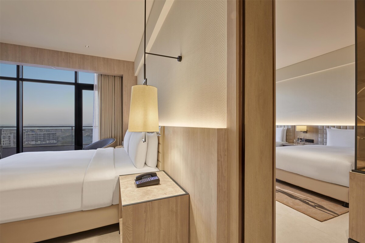 Une chambre d’hôtel avec un grand lit, une table de chevet avec une lampe et une vue sur la ville grâce à de grandes fenêtres.