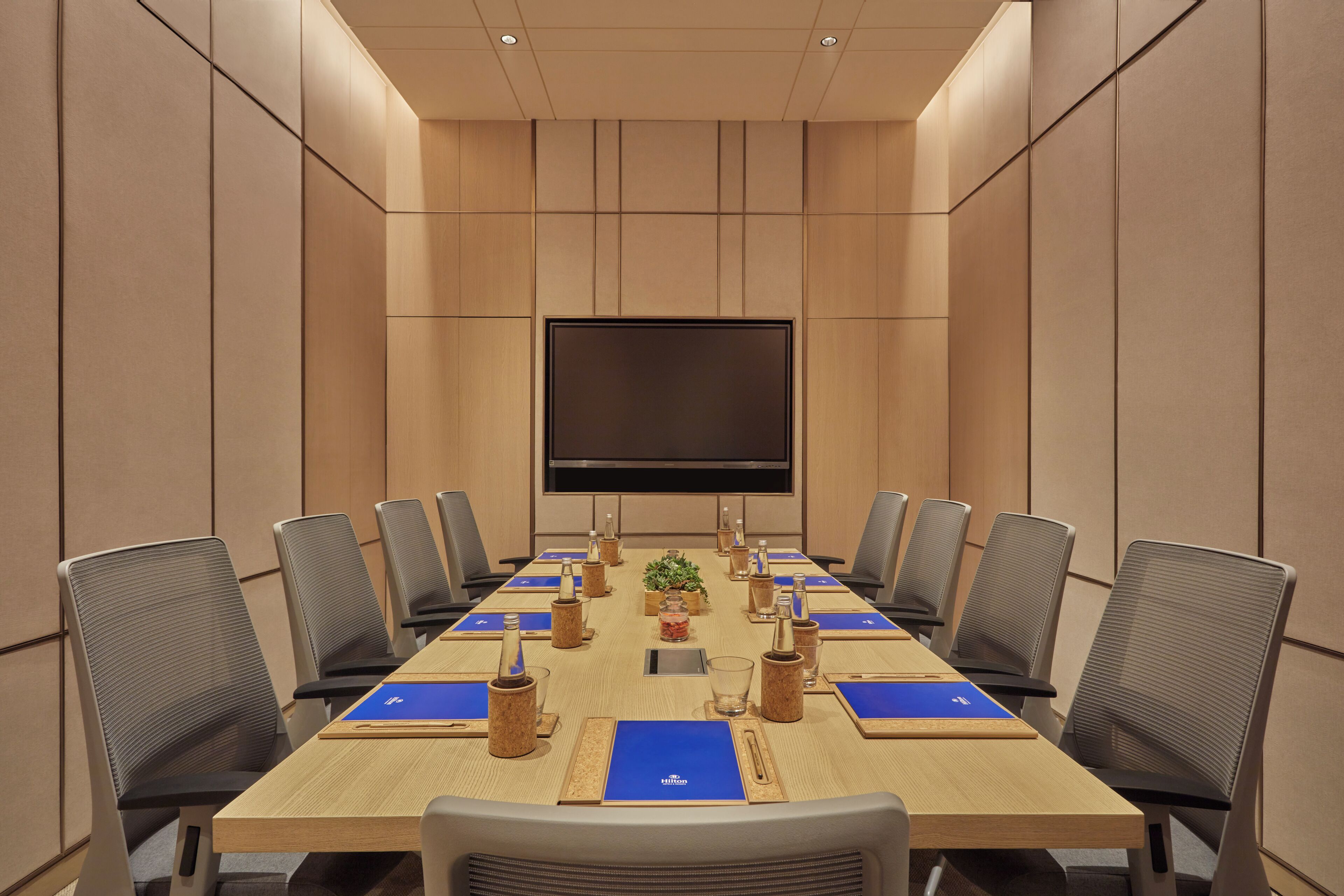 A conference room with a long table, chairs, a TV screen, and a plant.