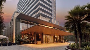 Entrada de hotel moderna con un área exterior techada, amplias ventanas y un jardín ajardinado con palmeras.