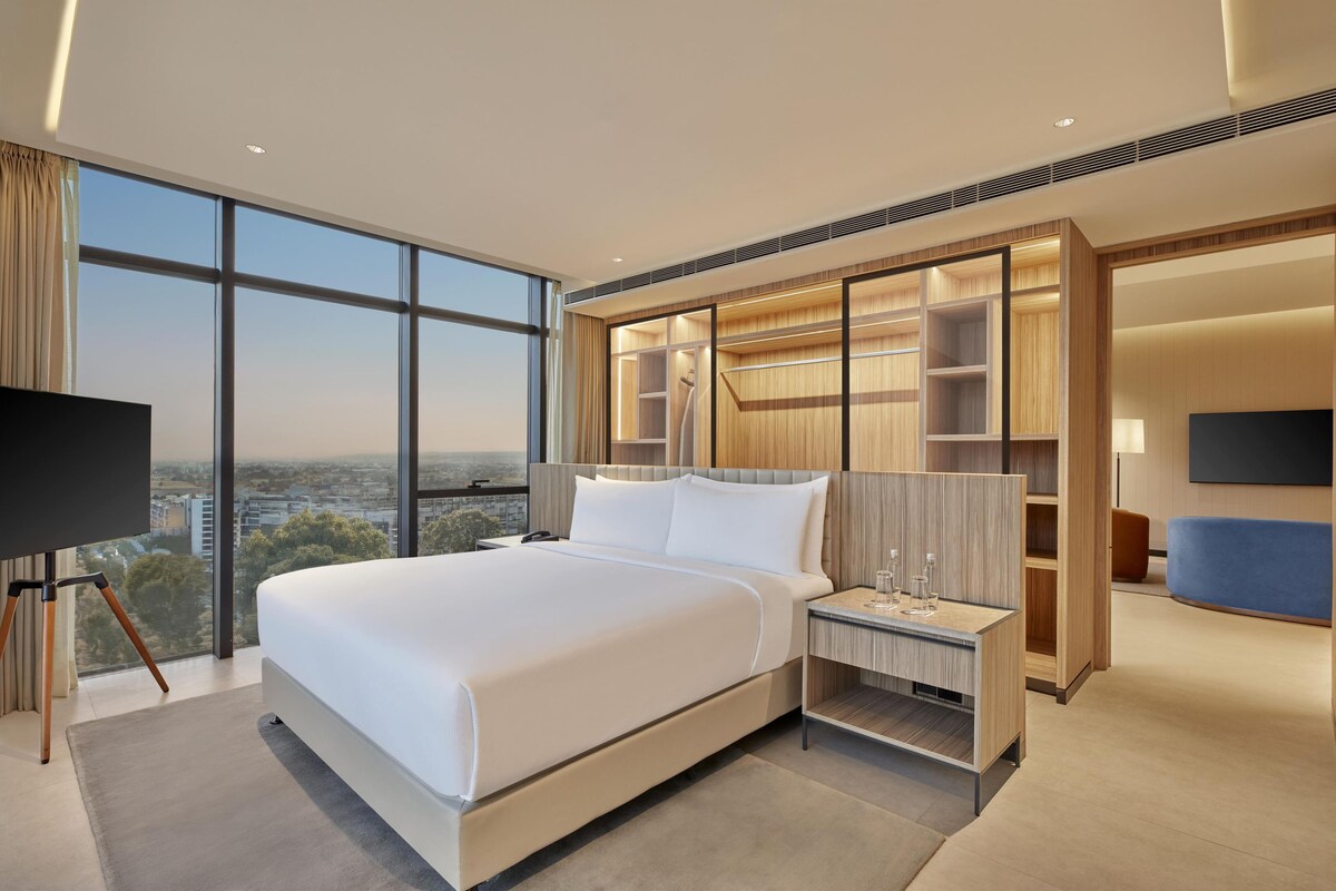 Une chambre d’hôtel moderne dotée d’un grand lit, d’une télévision, d’un bureau avec une lampe et offrant une vue sur la ville grâce à de grandes fenêtres.
