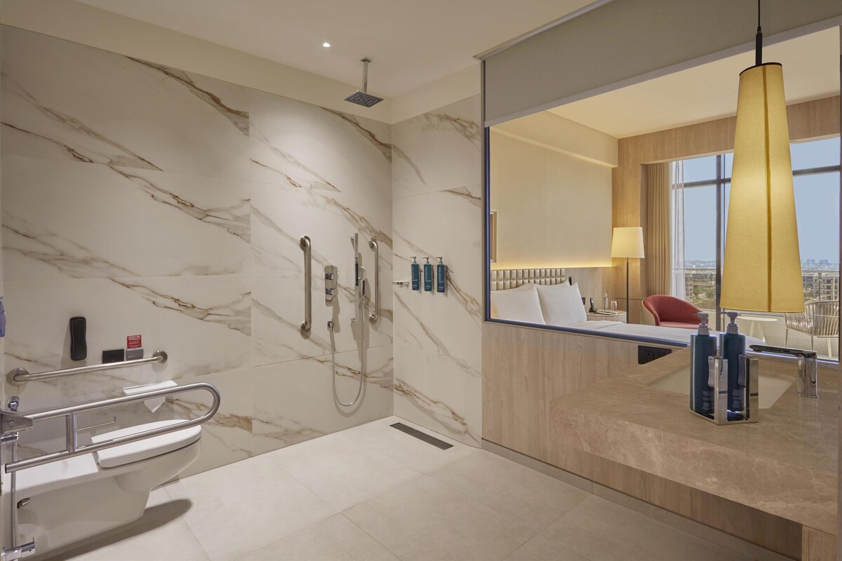 Une salle de bain moderne avec un mur en marbre, une douche, des toilettes et un lavabo. Un miroir reflète une pièce où l’on aperçoit une chaise et une lampe.