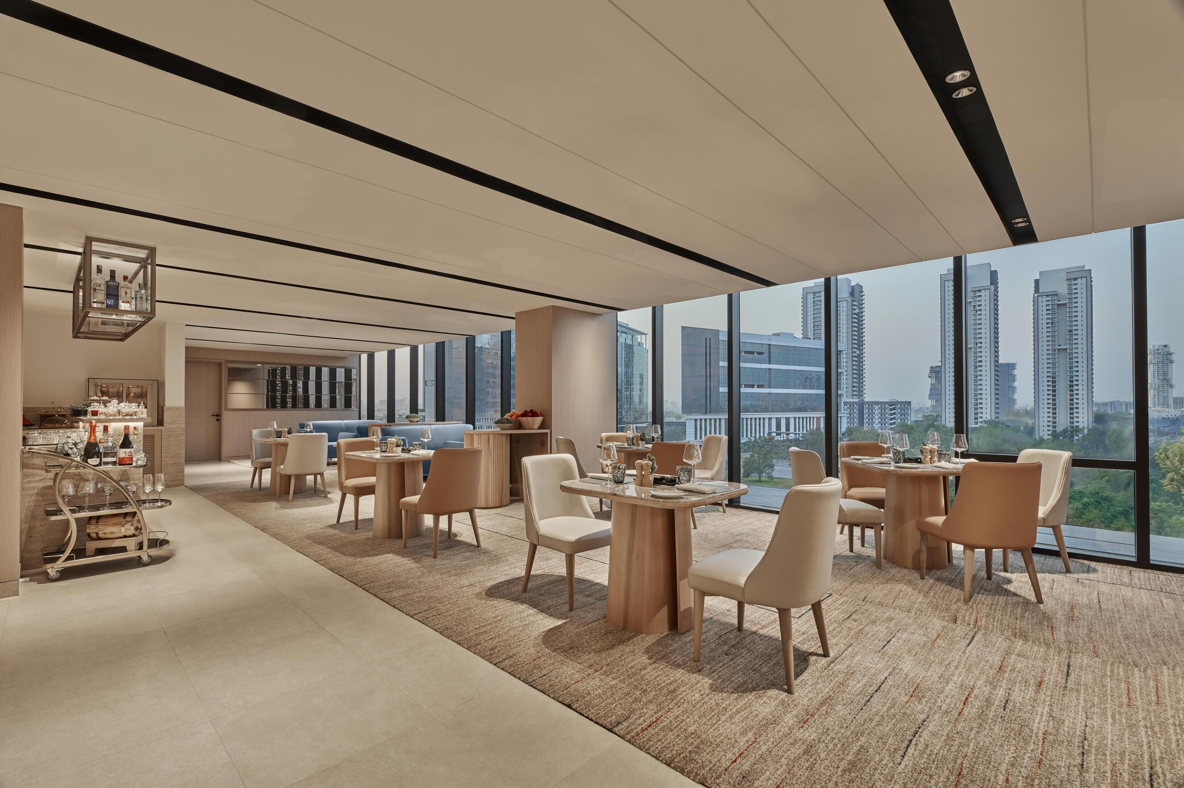 A modern hotel lobby with high ceilings, large windows offering city views, and a mix of seating arrangements including round tables and chairs.