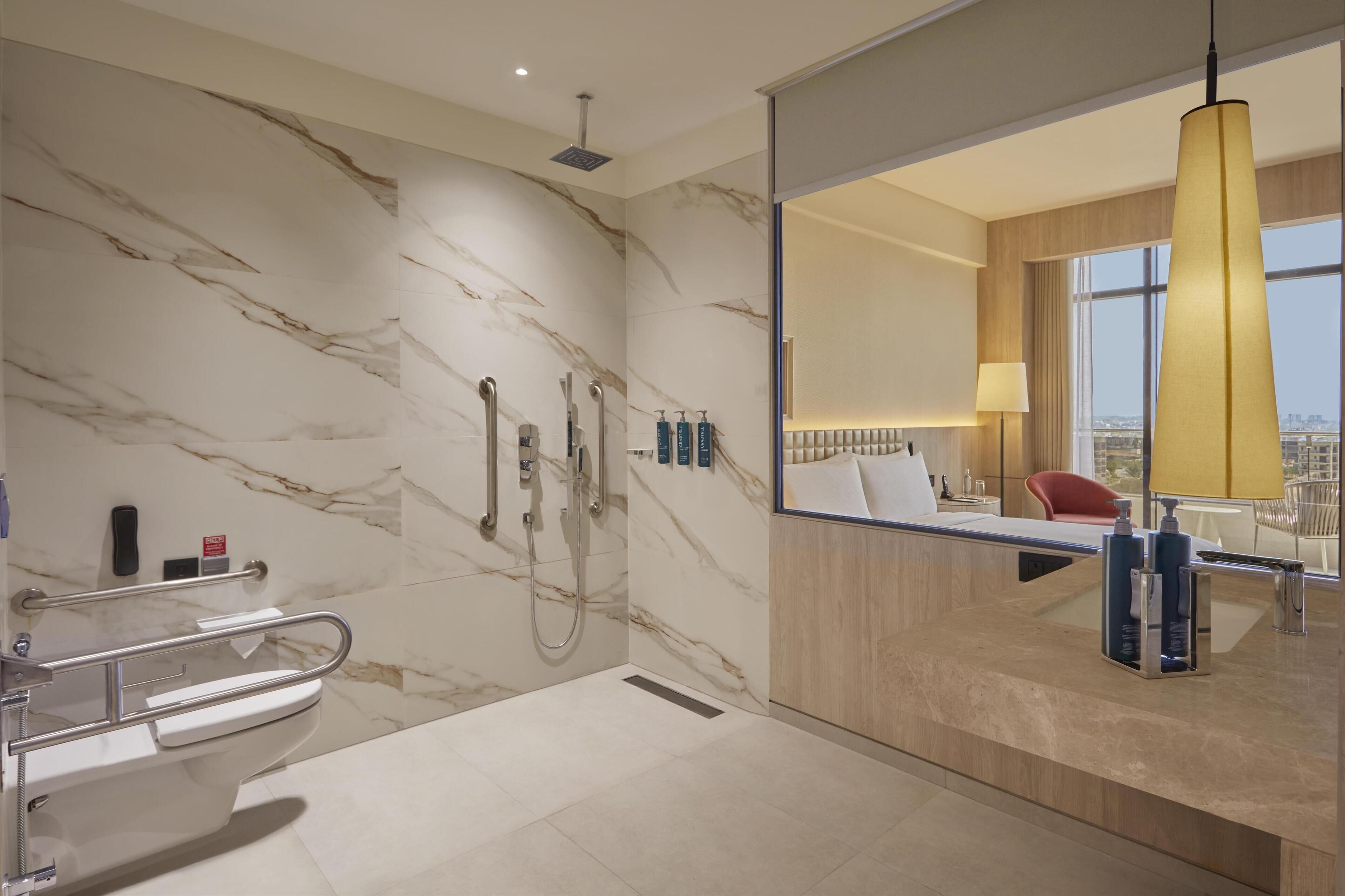Un baño moderno con pared de mármol, ducha, inodoro con barandilla y un espejo grande que refleja vistas de la ciudad.