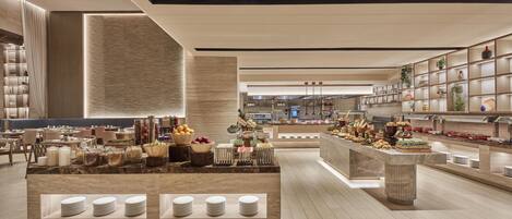 Un hall d’hôtel moderne avec un buffet central, des espaces aménagés pour s’asseoir et un comptoir de réception.