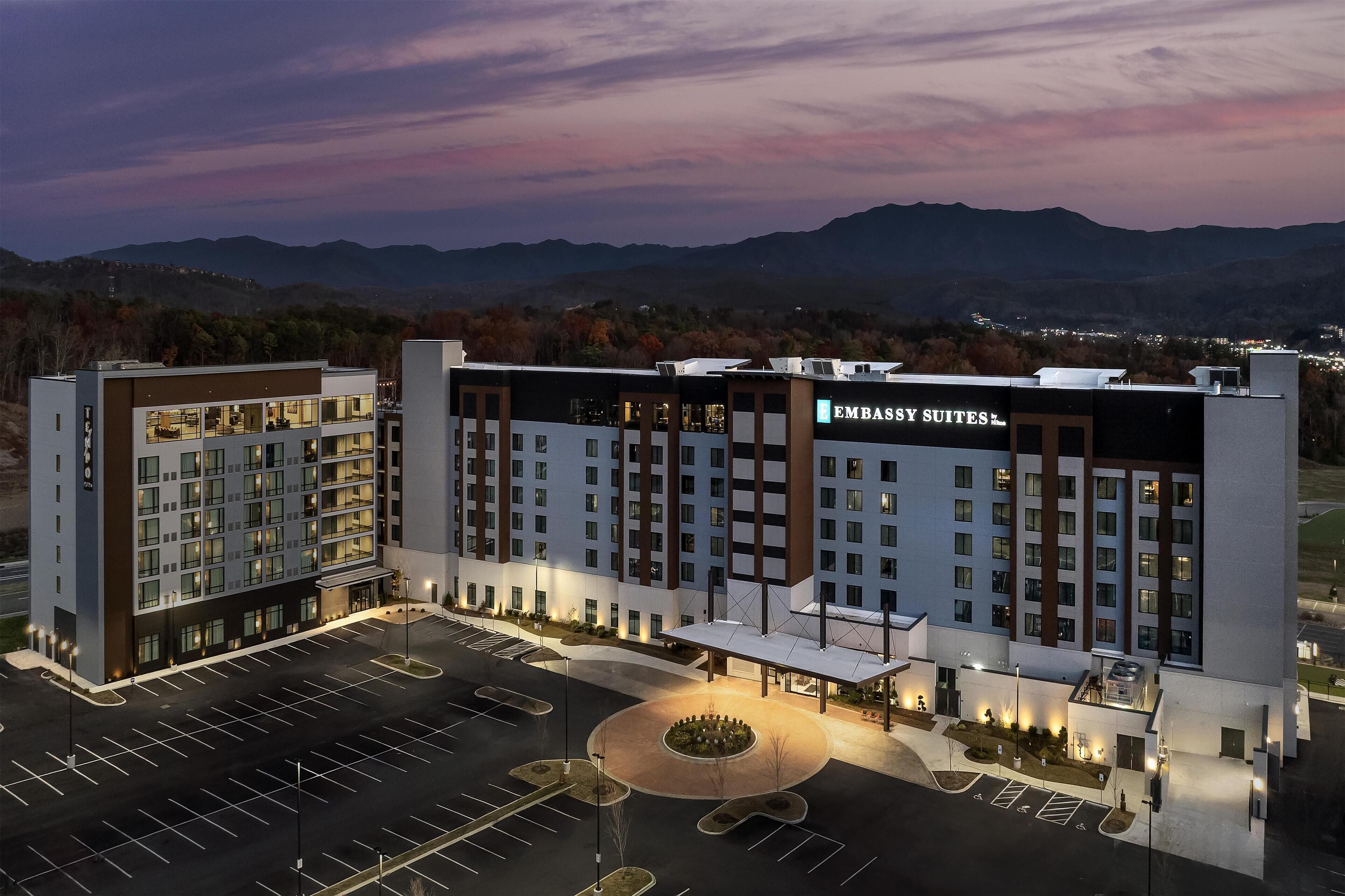 A modern hotel with multiple floors, a parking lot, and a view of mountains in the background.