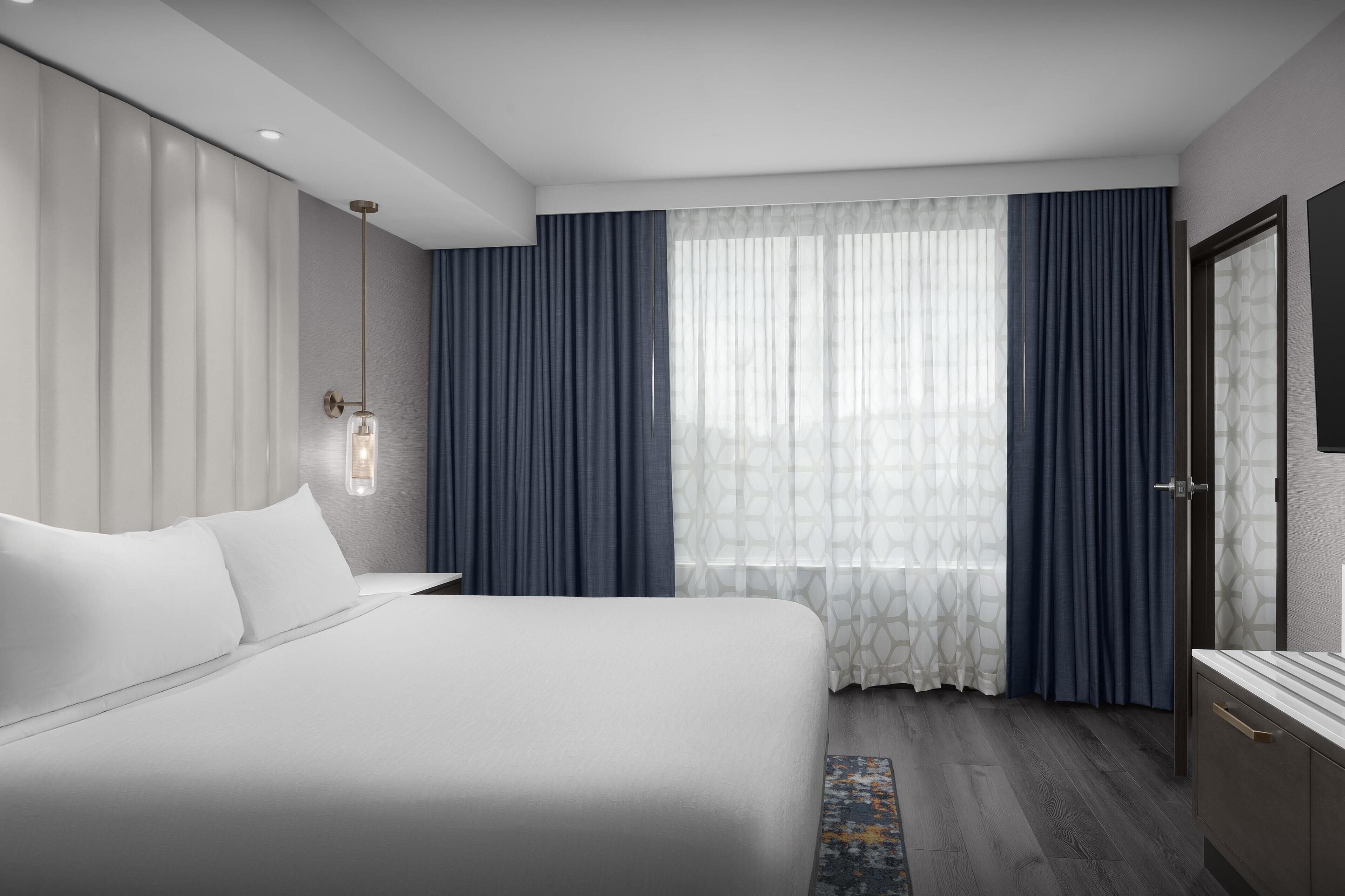 Une chambre d’hôtel moderne, dotée d’un grand lit, d’une table de chevet, d’une armoire et d’une grande fenêtre avec des rideaux.