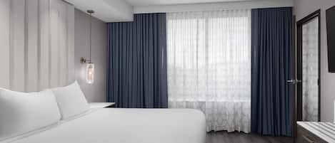 A modern hotel room with a large bed, a bedside table, a wardrobe, and a large window with curtains.
