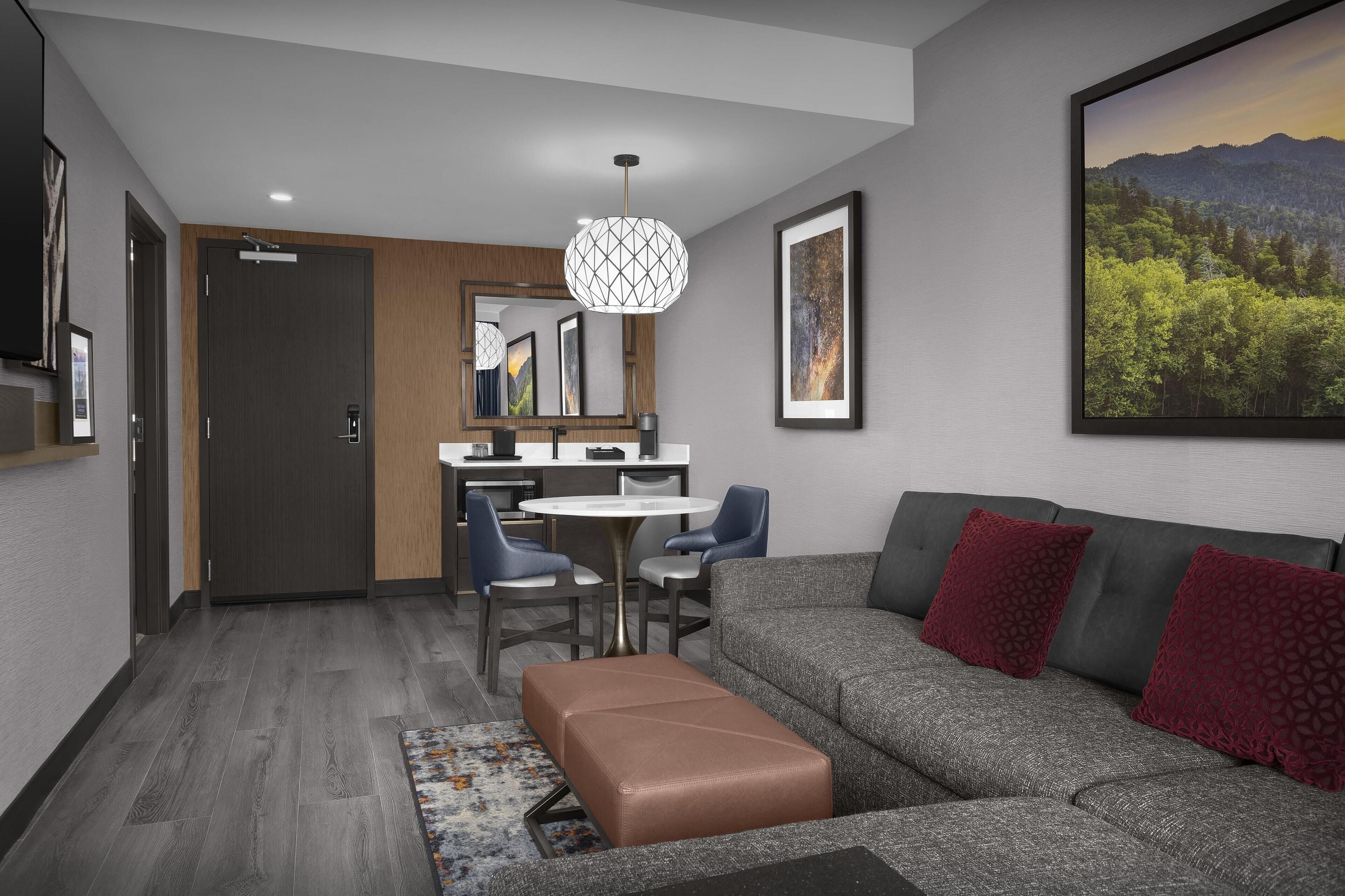 A modern living room with a grey sofa, red cushions, a brown ottoman, and a dining area with a round table and chairs.
