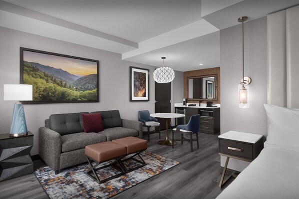 A modern hotel room with a grey sofa, a small table, a bench, a desk, and a bed.