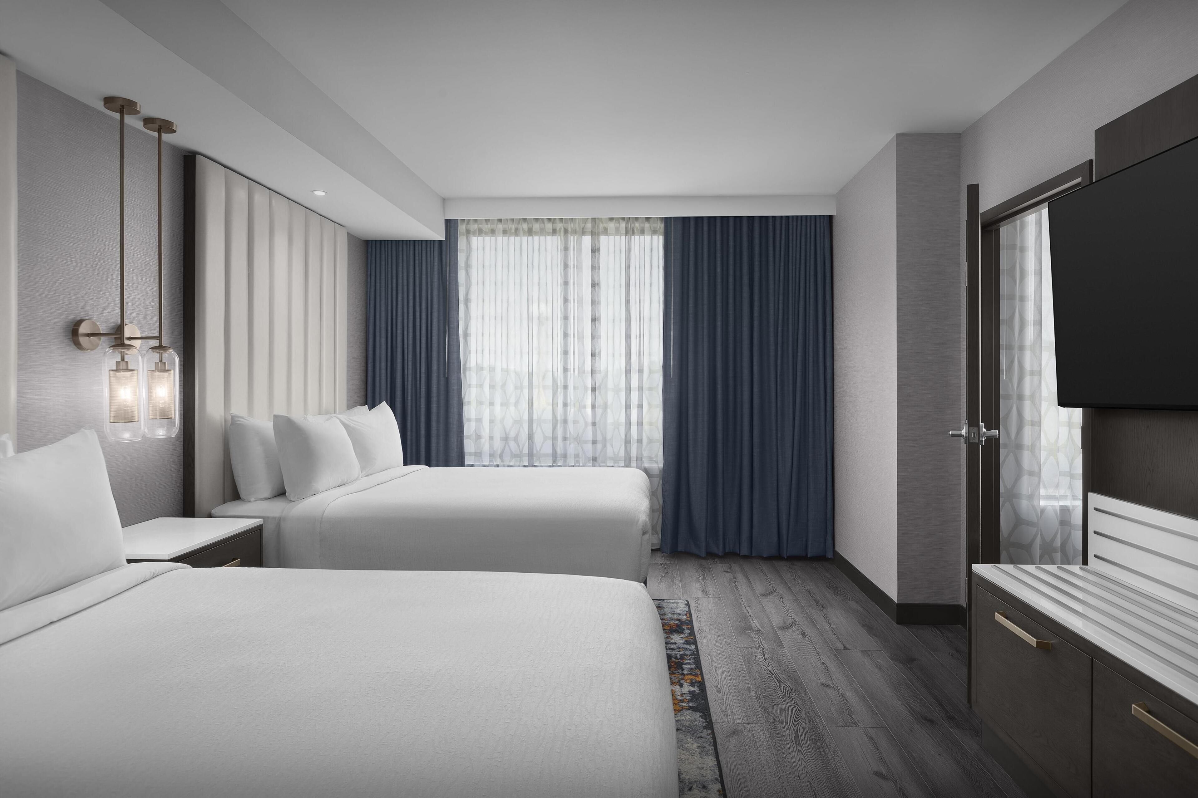 Habitación de hotel moderna con dos camas, un ventanal con cortinas, un televisor de pantalla plana y suelo de madera.