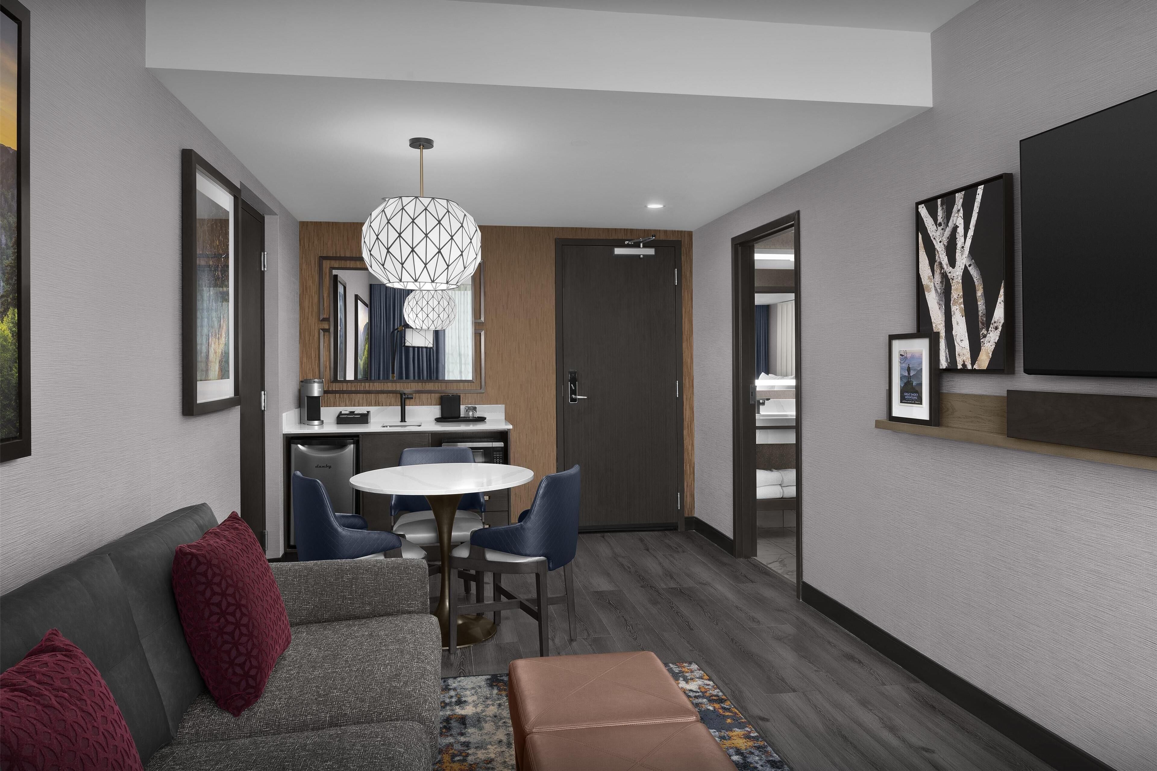 Una habitación de hotel moderna con un sofá gris, una mesa de comedor redonda y una kitchenette.