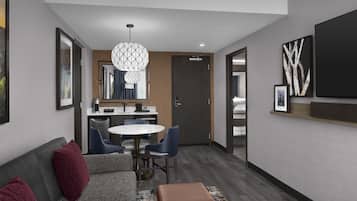 A modern hotel room with a gray sofa, a round dining table, and a kitchenette.