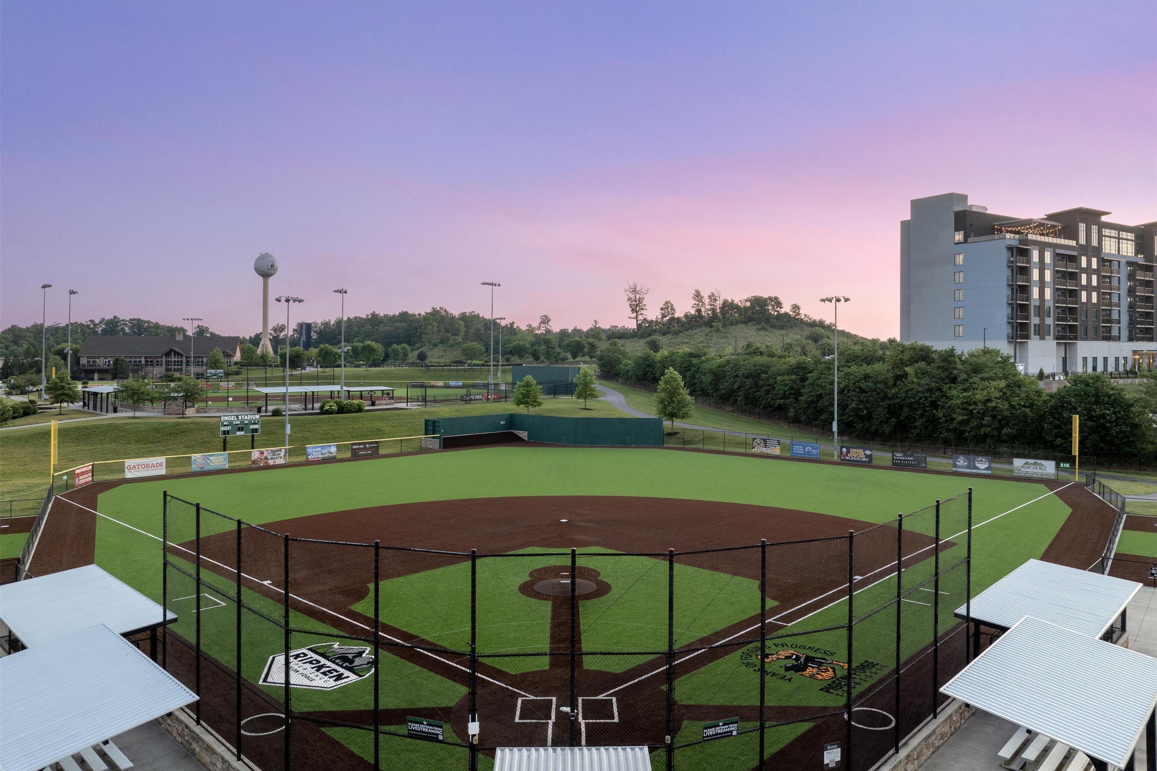 A baseball field with a large scoreboard, a tall observation tower, and a modern hotel in the background.