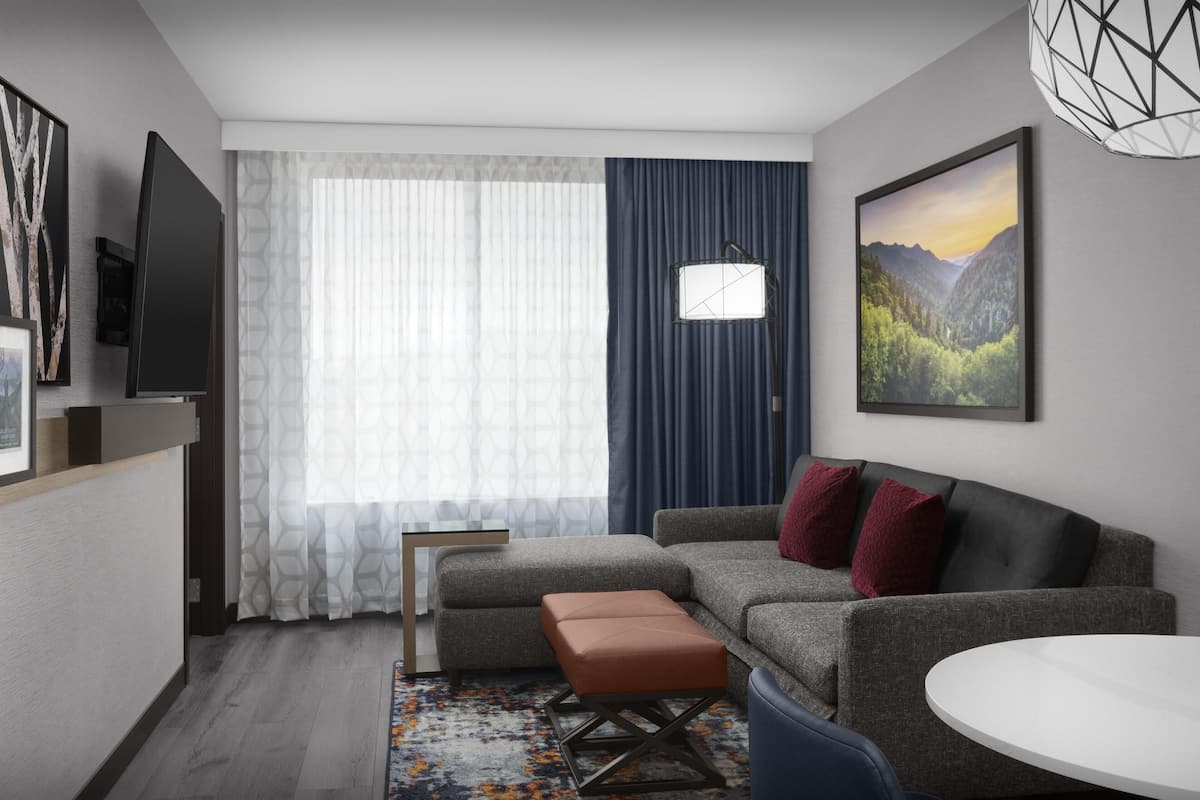 Een moderne woonkamer met een grijze bank, een bruin voetenbankje, een witte ronde tafel en een groot raam met dunne gordijnen.