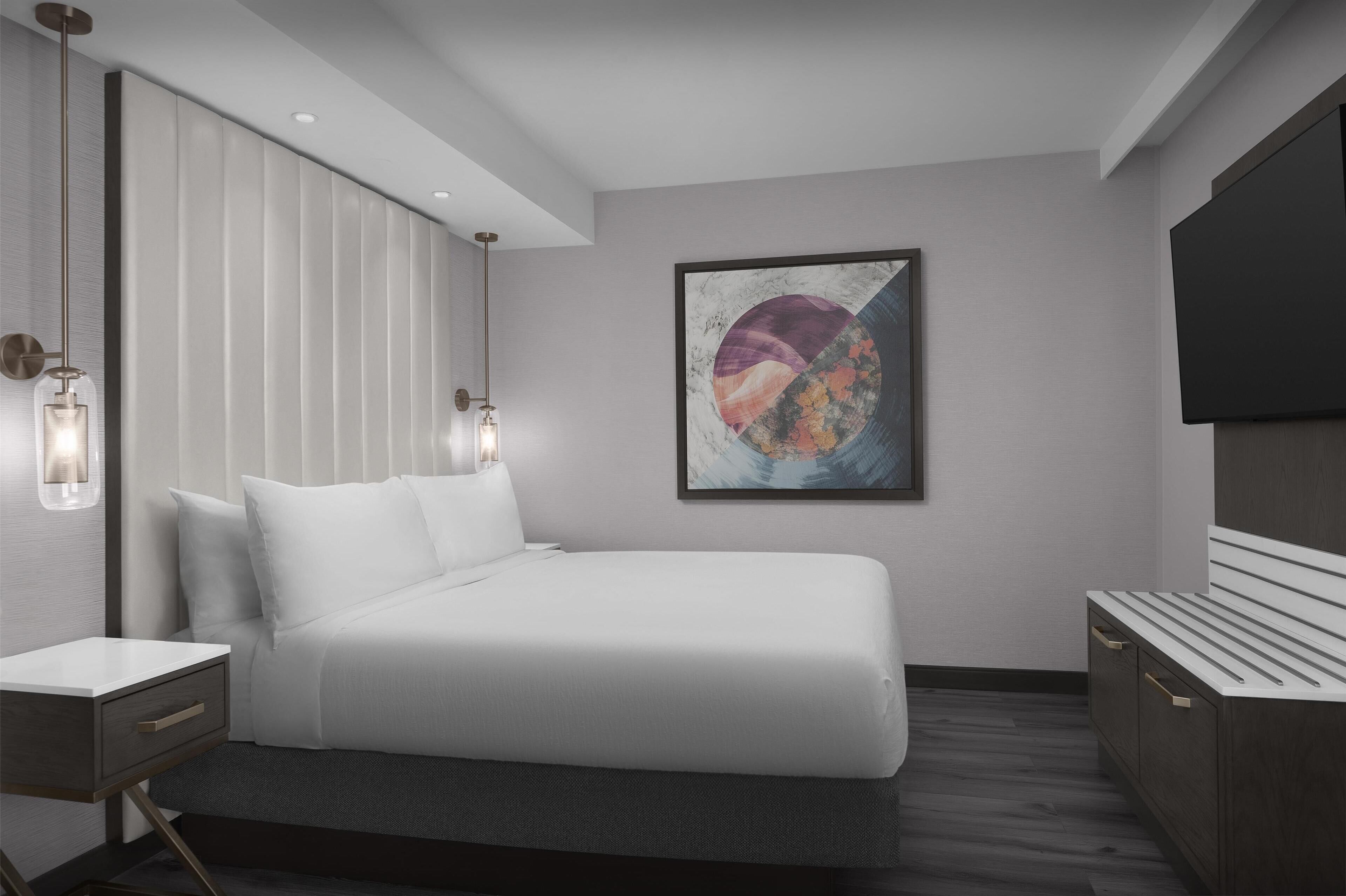 Une chambre d’hôtel moderne avec un lit soigneusement fait, une table de chevet avec une lampe, un grand tableau abstrait accroché au mur et un téléviseur à écran plat fixé au mur.