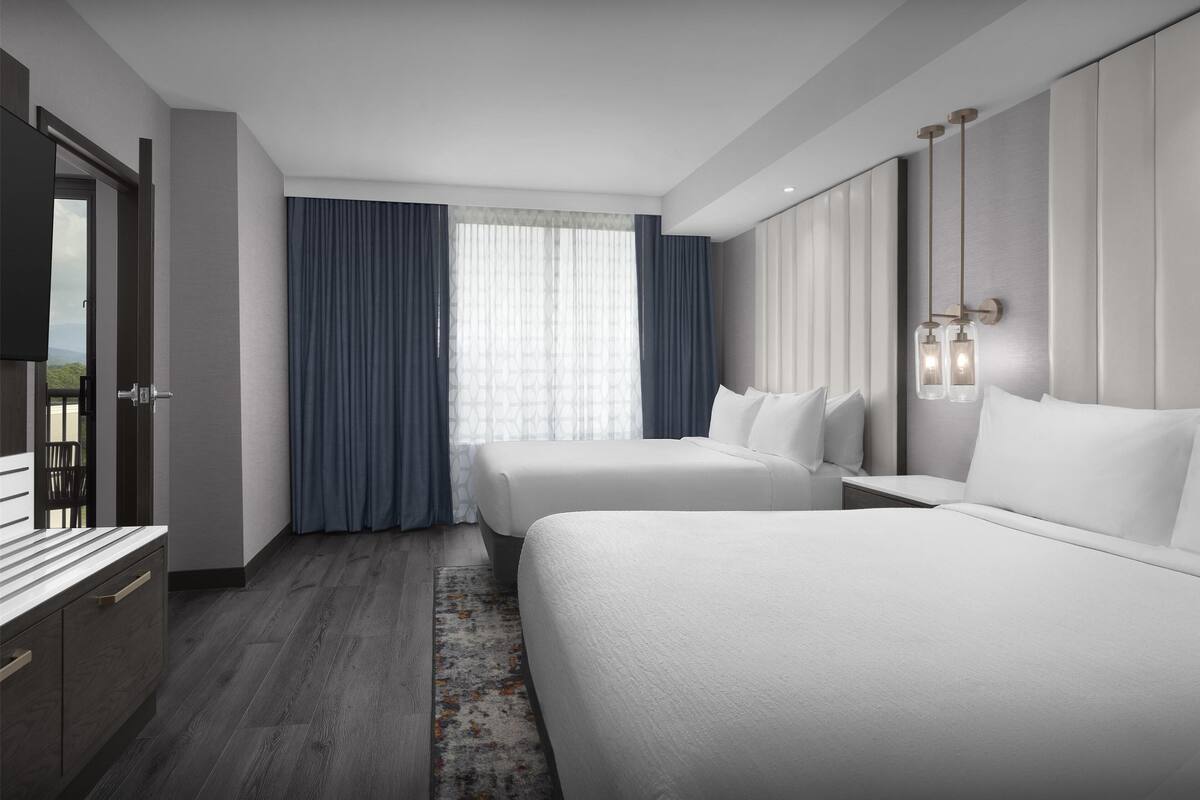 Een moderne hotelkamer met twee bedden, een groot raam met gordijnen, een flatscreen televisie en een houten vloer.