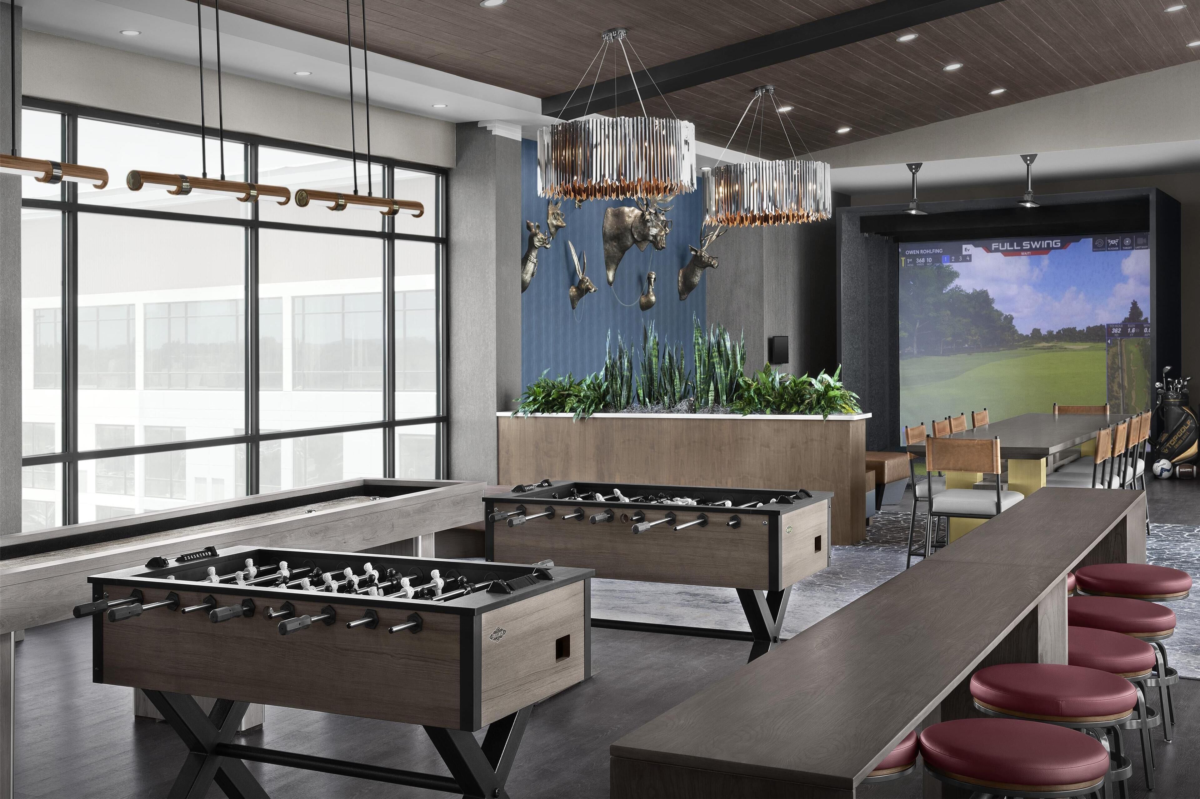 Un espacio interior moderno con dos mesas de fútbol de mesa, una larga mesa de comedor con sillas rojas acolchadas y una pantalla grande que muestra un partido de golf.