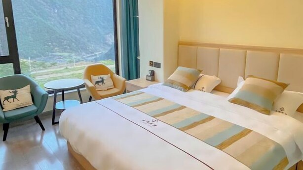 Habitación de hotel con una cama grande, una mesita, dos sillas y vistas a montañas a través de un ventanal amplio.