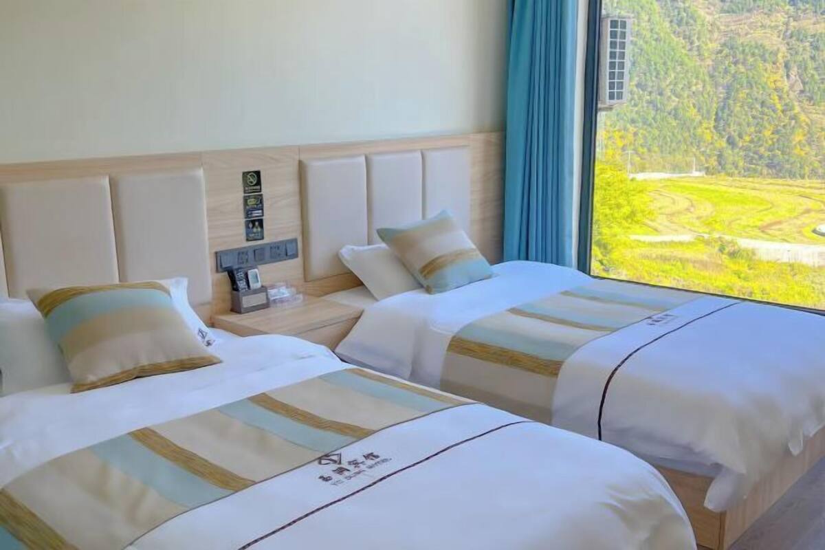 Een hotelkamer met twee bedden, een nachtkastje, een raam met uitzicht op een groen landschap en een houten hoofdeinde.
