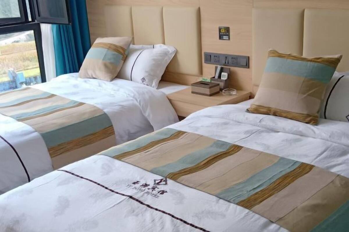 Een hotelkamer met twee bedden, elk voorzien van gestreept beddengoed en een hoofdbord. Er staat een nachtkastje met een lamp en een klein doosje, mogelijk voor toiletartikelen.