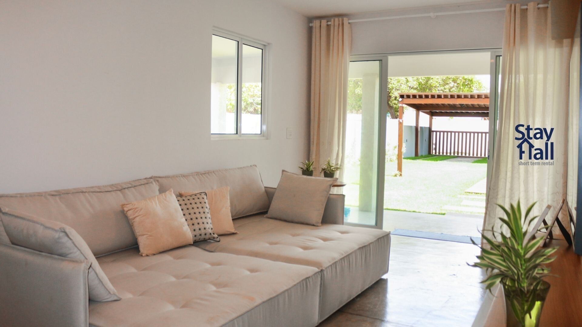 Uma sala de estar moderna com um sofá bege, duas almofadas e uma pequena mesa com plantas em vasos. Uma porta de vidro deslizante dá acesso a uma área externa com pérgola de madeira.