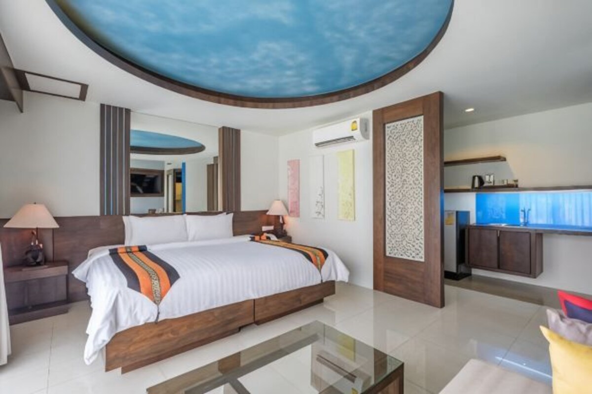 A modern hotel room with a large bed, a glass coffee table, a kitchen area, and a unique ceiling design.
